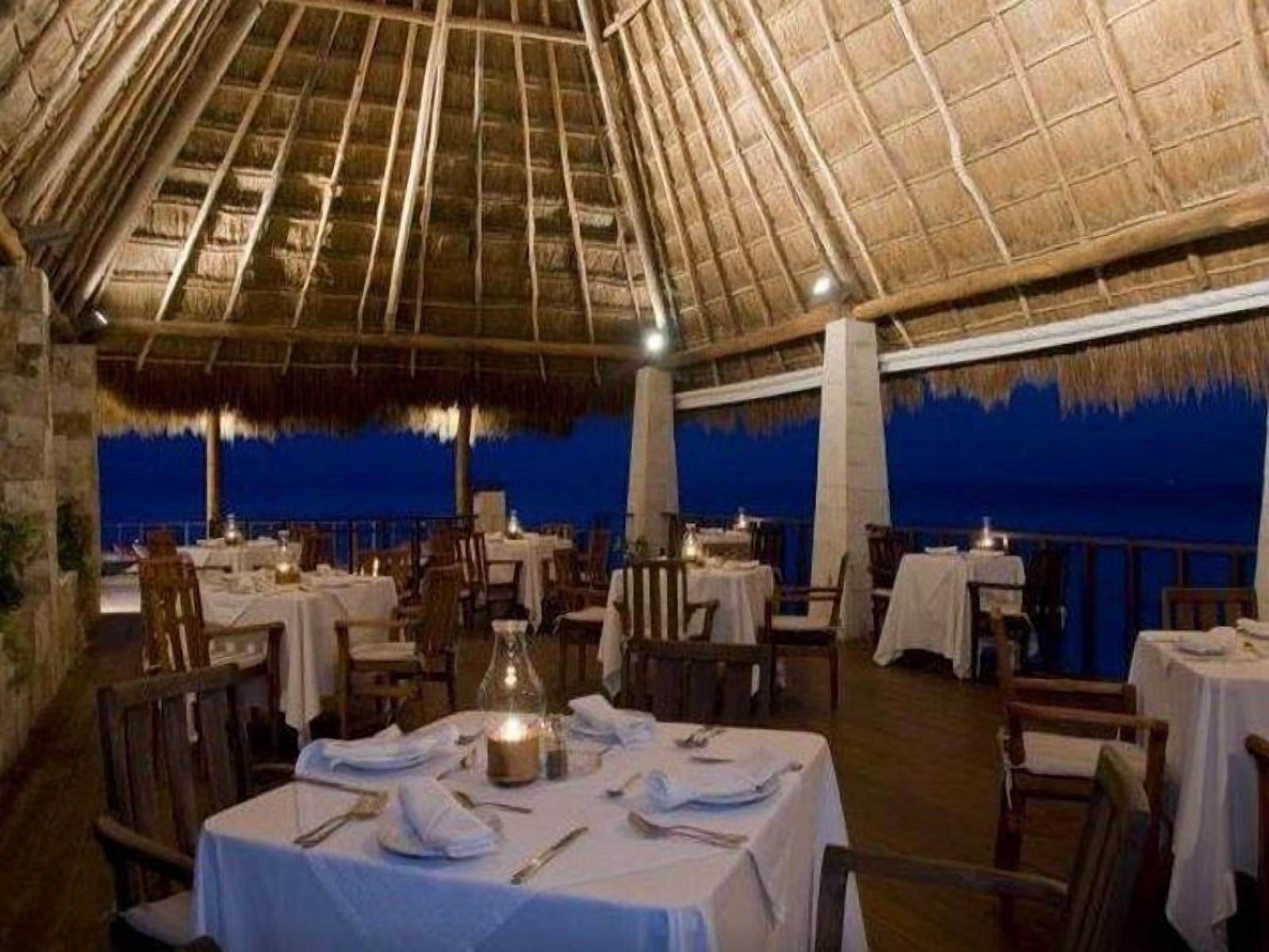Ein Essbereich im Freien mit reetgedeckten Dächern, gedeckten Tischen und Blick auf das Meer in der Abenddämmerung.