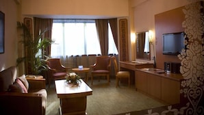 Habitación de hotel con sofá, dos sillones, una mesita con un florero, televisor y un ventanal con cortinas.