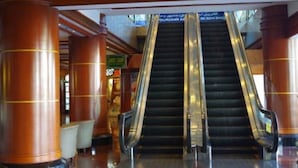 Una escalera mecánica en un edificio comercial con columnas de madera y un cartel que indica una zona de tiendas.