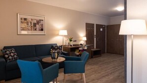 A modern living room with a dark blue sofa, two matching armchairs, a small round table with a lamp, and a wooden floor. - Melantrich Sivek Hotels (Prague)
