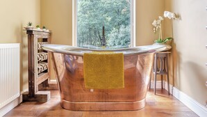 A bathroom with a freestanding copper bathtub, a window with a view of trees, and a small wooden table with plants.