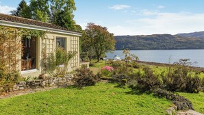 A quaint cottage with a stone wall and a window overlooking a lake, surrounded by lush greenery and a clear sky.
