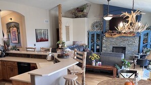 A spacious kitchen with a central island, wooden chairs, a dining table with a floral tablecloth, and a stone fireplace. - Mountain View Spacious Ranch Home AMAZING VIEW (Milliken)