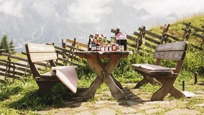 Ein rustikales Freigelände mit einem Holztisch und zwei Bänken, umgeben von einem Holzzaun und einer Berglandschaft im Hintergrund.