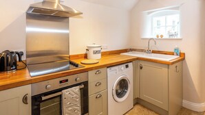 A compact kitchen with a stainless steel range hood, a built-in oven, a washing machine, and a window with a view of buildings. - 1 bedroom accommodation in Thirlby, near Thirsk (Thirlby, near Thirsk)