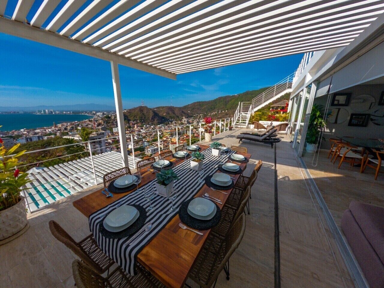 A rooftop dining area with a long table set for six, overlooking a cityscape and mountains.