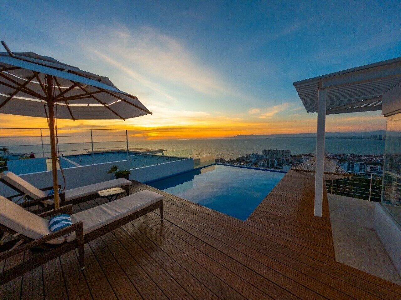 A rooftop infinity pool with a wooden deck, lounge chairs, and a pergola.