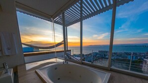 Une salle de bain moderne avec une grande baignoire, un lavabo et une vue sur la ville et la mer au coucher du soleil.