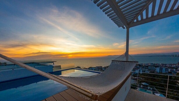 Un hamac sur une terrasse en bois donnant sur une piscine et offrant une vue sur le coucher du soleil.