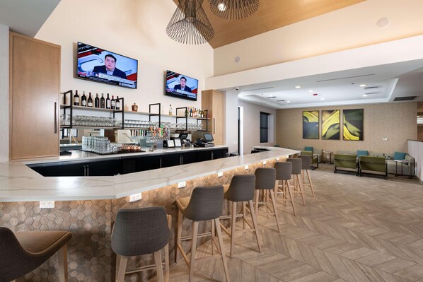 A bar area with a long counter, multiple bar stools, and two televisions mounted on the wall.