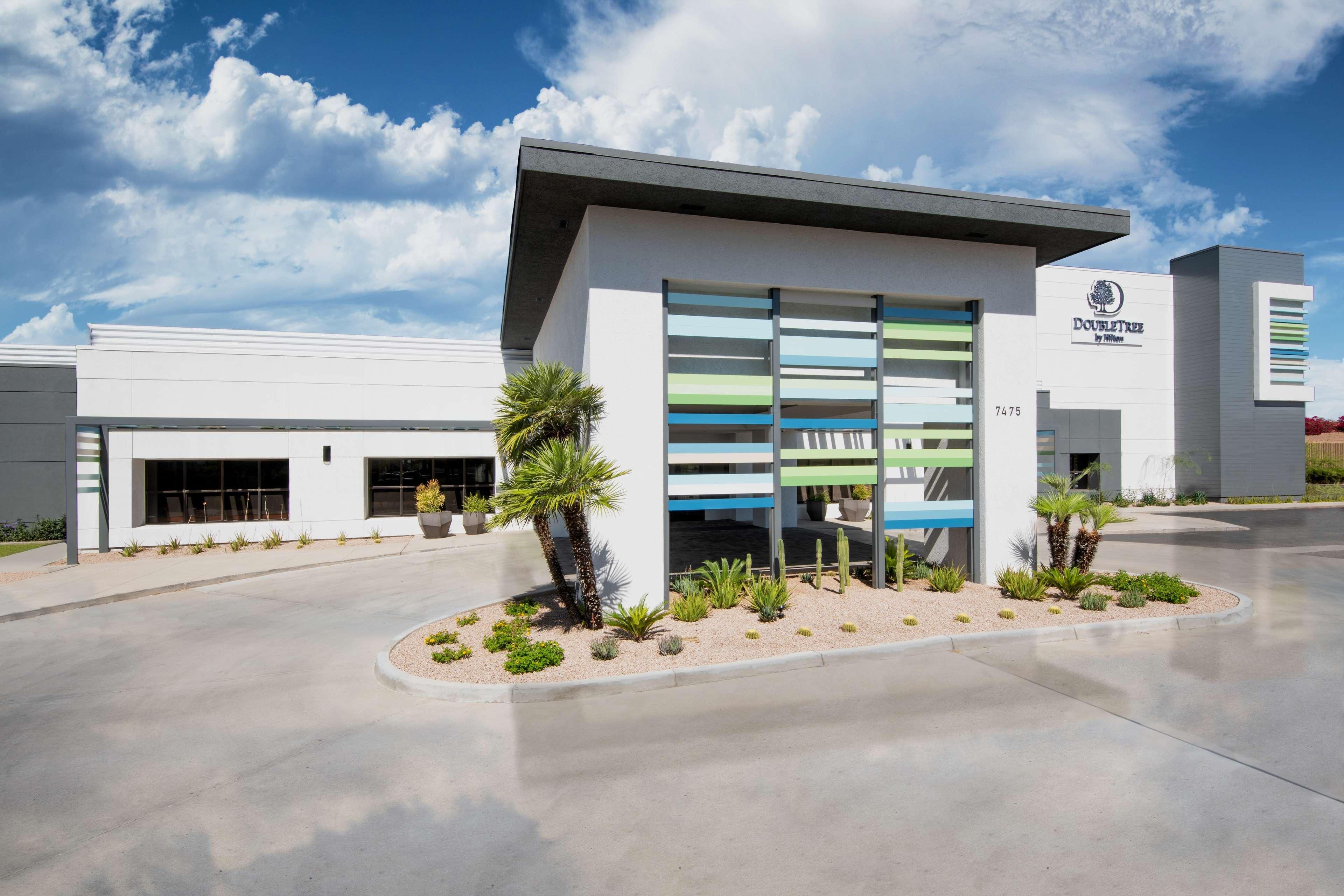 A modern building with a flat roof, a sign reading 'Doubleday by Hilton,' and a parking lot with palm trees and shrubs.