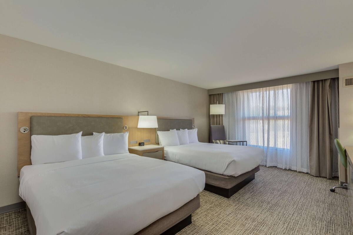 A hotel room with two beds, a desk, a chair, and a large window with curtains.