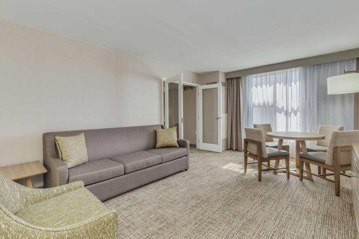 A hotel room with a sofa, a dining table with chairs, a small side table, and a large window with curtains.