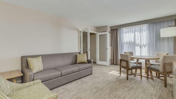 Habitación de hotel con sofá, mesa de comedor con sillas, una mesita auxiliar y un ventanal con cortinas.