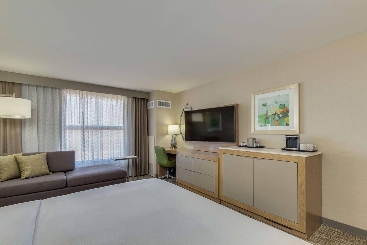 A hotel room with a bed, a sofa, a desk, a television, and a painting on the wall.