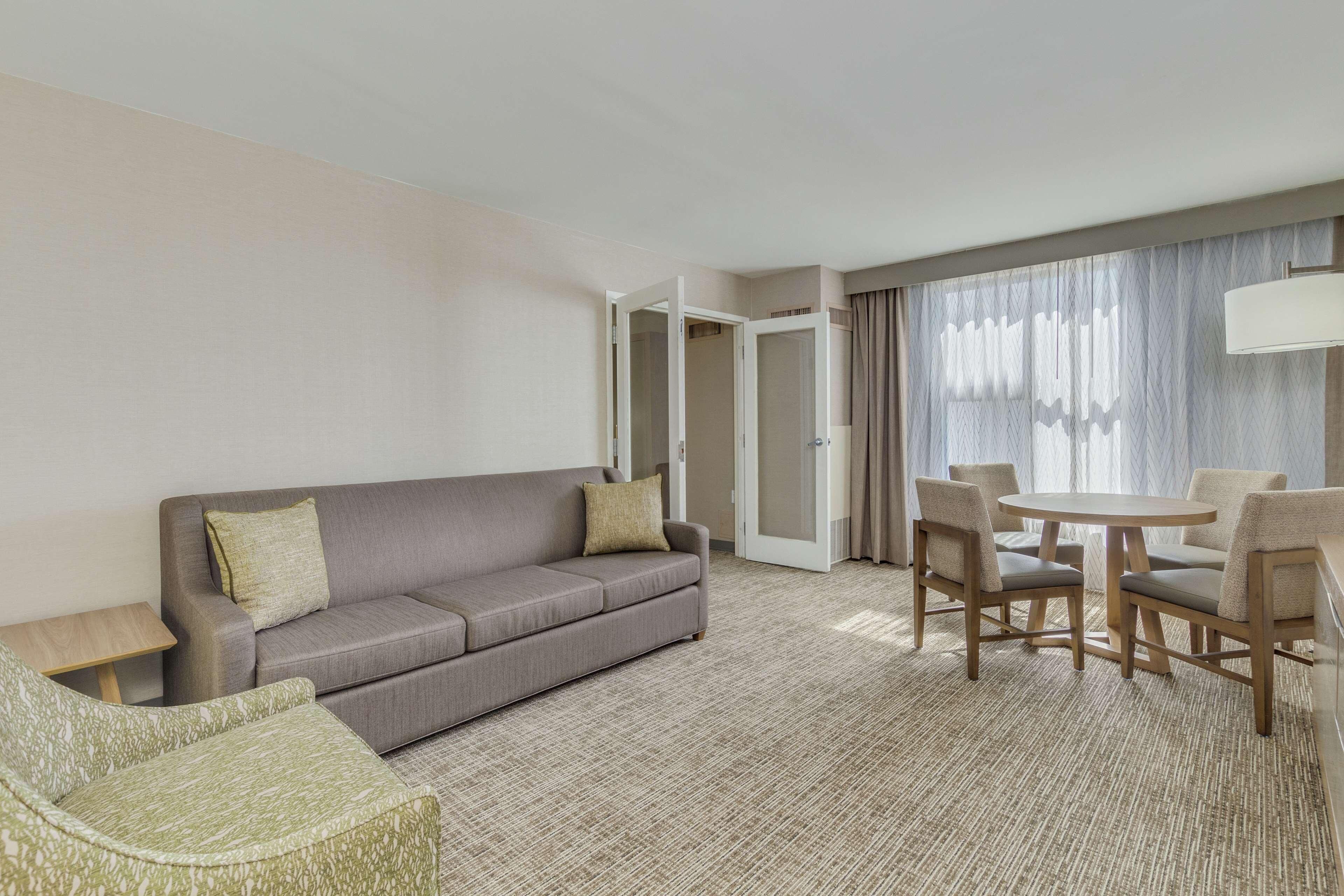 Habitación de hotel con sofá, mesa de comedor con sillas, una mesita auxiliar y un ventanal con cortinas.