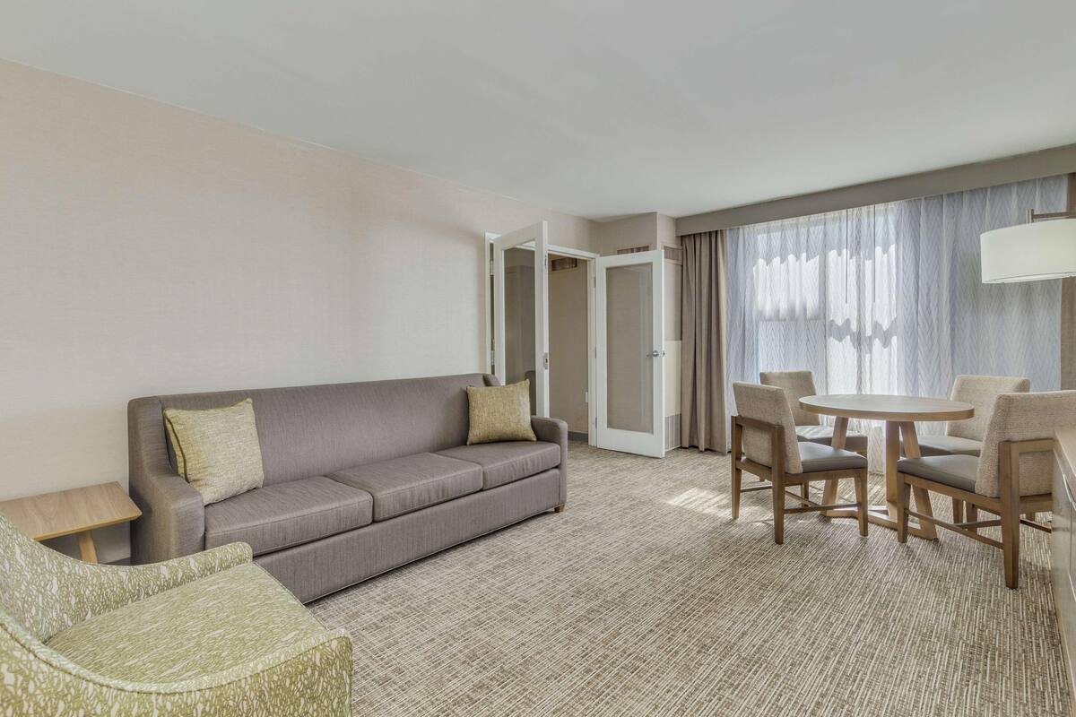 A hotel room with a sofa, a dining table with chairs, a small side table, and a large window with curtains.