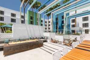 Eine moderne Lounge-Bereich im Freien mit Feuerstelle, Sitzgelegenheiten und Blick auf ein Gebäude mit zahlreichen Fenstern und Balkonen.