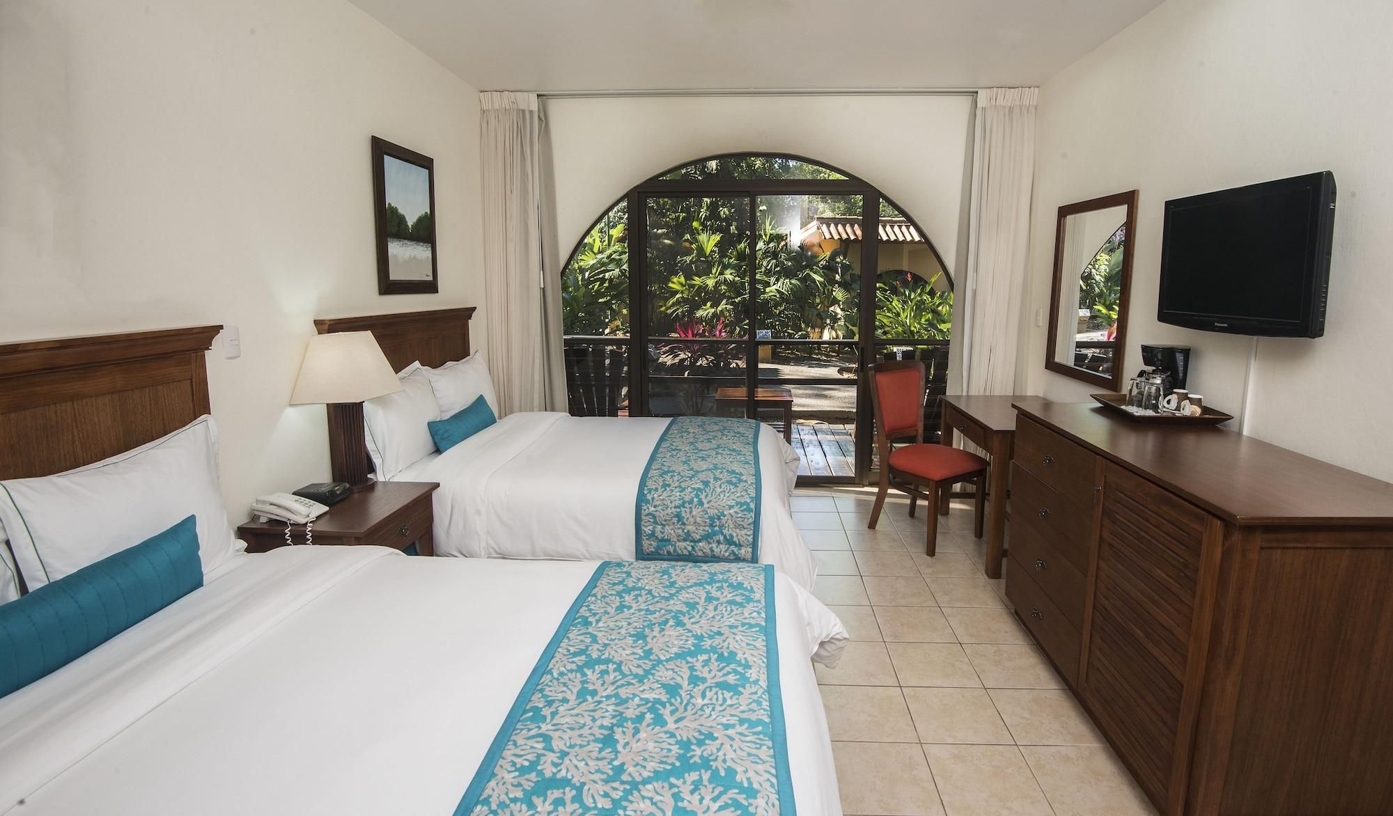 A hotel room with two beds, a desk, a chair, a television, and a balcony with a view of greenery.
