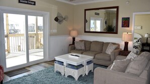 A cozy living room with a beige sofa, a white coffee table, and a blue rug. There is a wooden deck visible through the glass door. - Island Time II - 102 Raphael S (Dauphin Island)
