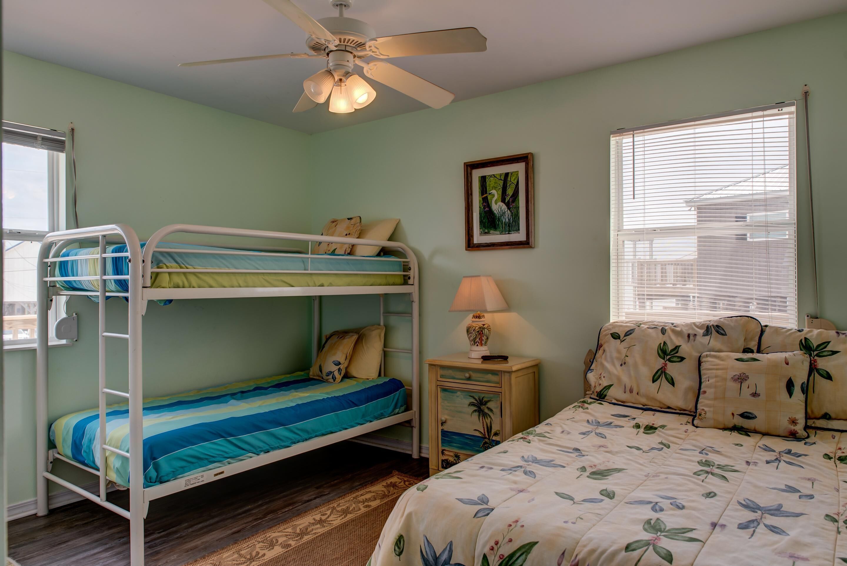 A bunk bed room with a ceiling fan, a bedside table with a lamp, and a window with blinds.