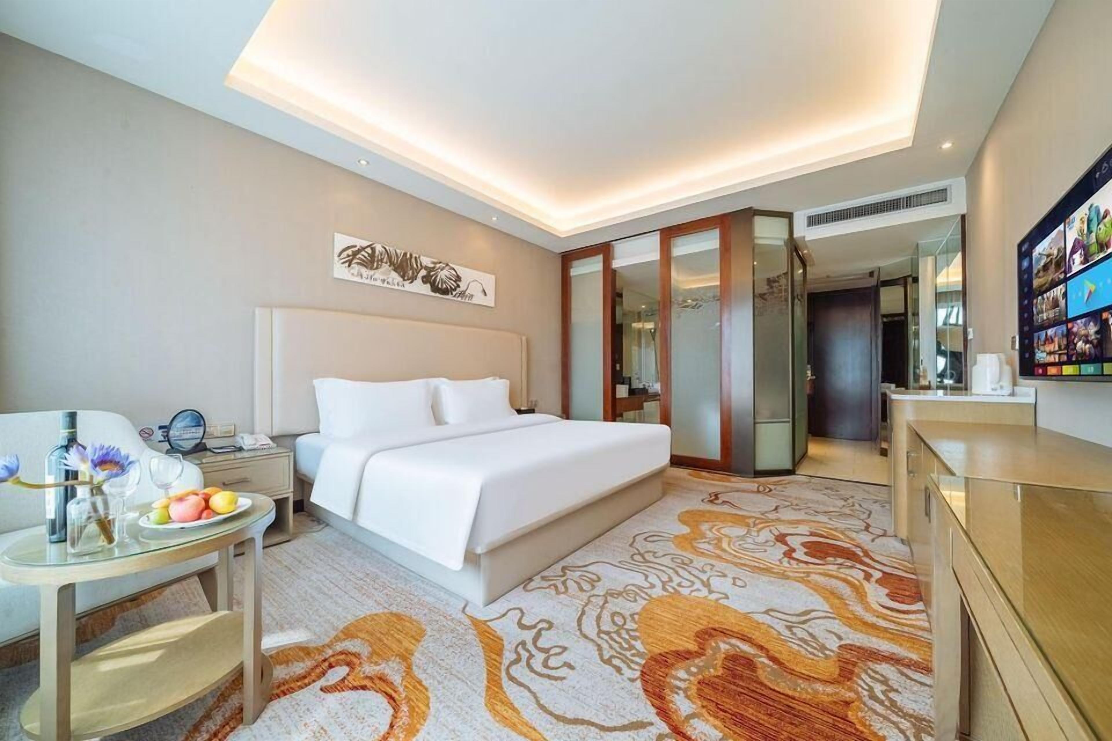 A hotel room with a large bed, bedside table with a lamp, a small table with fruits and a vase, and a TV on the wall.
