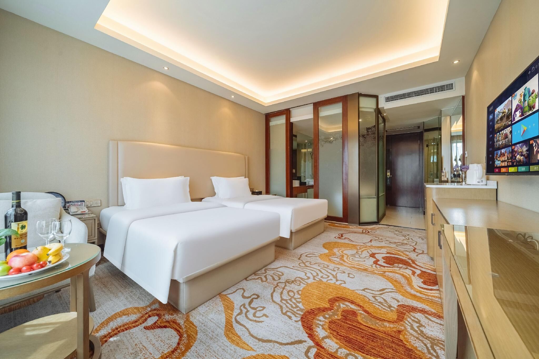 A hotel room with a double bed, a small table with fruit and a bottle, a mirror, and a TV on the wall.