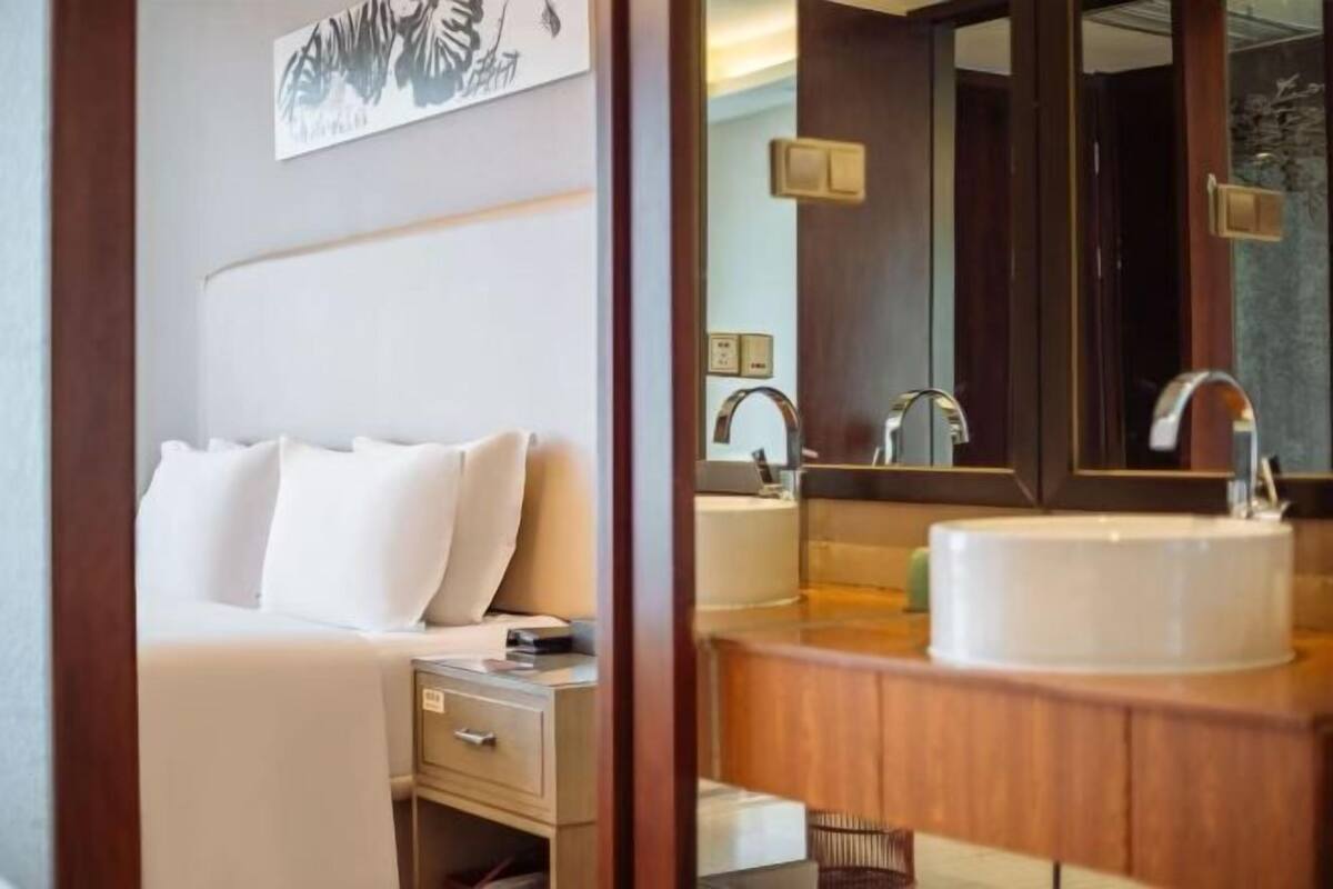 Une chambre d’hôtel avec un lit, une table de chevet et un grand miroir qui reflète une salle de bain avec un lavabo et un miroir.