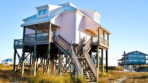A house with a balcony and stairs leading up to it, surrounded by grass and other houses. - Second Wind - 2612 Bienville Blvd (Dauphin Island)