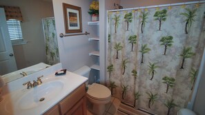 A bathroom with a white sink, a toilet, a shower curtain with palm tree patterns, and a framed picture on the wall. - Second Wind - 2612 Bienville Blvd (Dauphin Island)