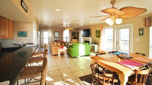 A spacious kitchen with a large island, dining area with chairs, a green sofa, and a television in the living room. - Second Wind - 2612 Bienville Blvd (Dauphin Island)