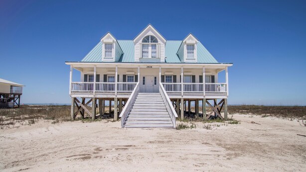 Una casa de playa de dos pisos con un balcón, una escalera que lleva a la entrada y una vista despejada de la playa.