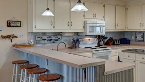 A kitchen with white cabinets, a central island with stools, a microwave, and a stove. - Water's Edge - 2227 Mallard (Dauphin Island)