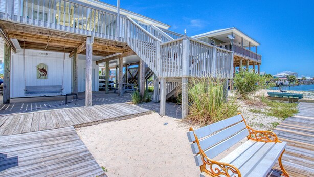 A beachfront property with a wooden deck, a covered seating area, and a staircase leading to a balcony. - Water's Edge - 2227 Mallard (Dauphin Island)