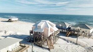 A beachfront property with multiple elevated structures, including a central building with a sloped roof and several smaller buildings with flat roofs, all supported by wooden stilts. - Kowabunga - 102 Shell Ct (Dauphin Island)