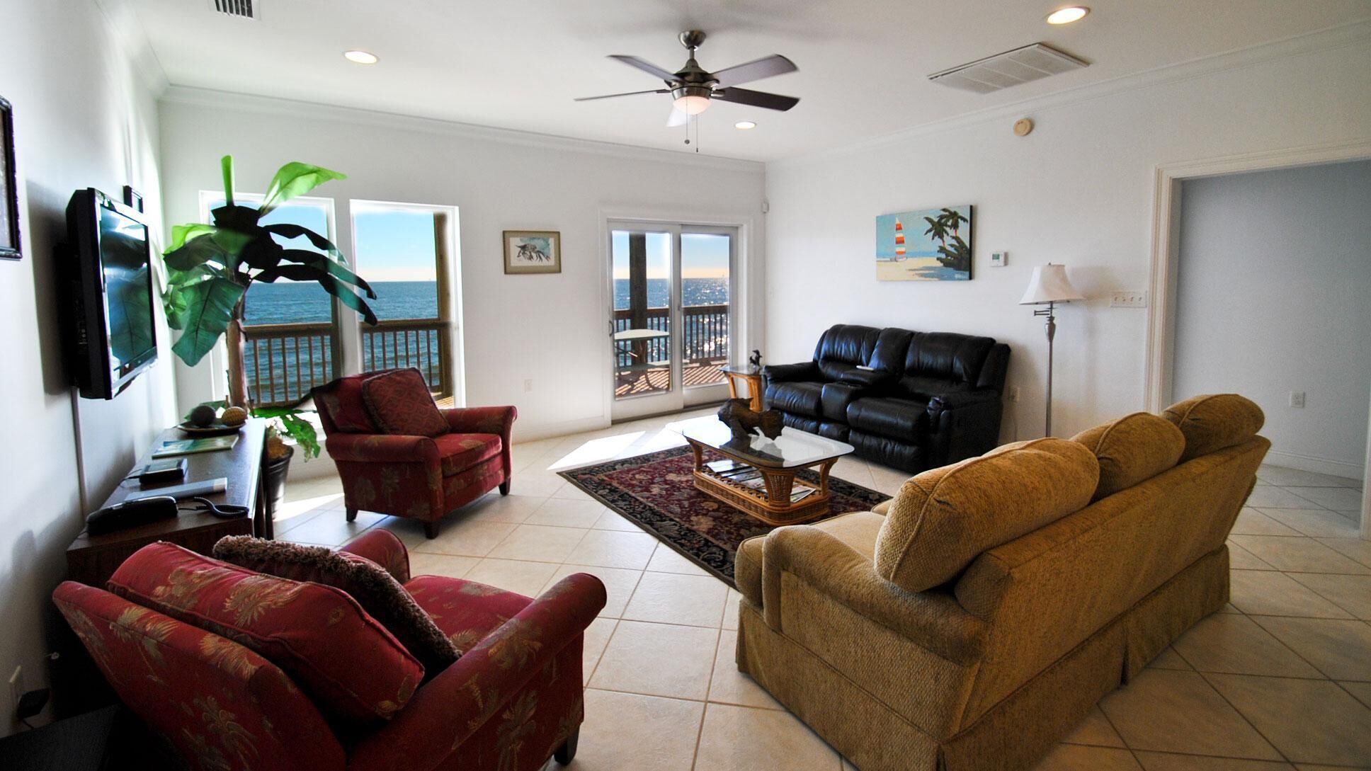 A spacious living room with a black leather sofa, a red armchair, and a brown sofa. There is a large window offering a view of the ocean.