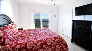 A bedroom with a bed, a dresser, a TV, and a balcony with a view of the sea.