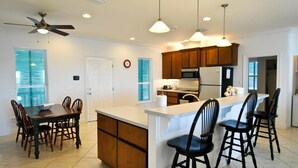 A modern kitchen with a central island, wooden cabinets, and a dining area with a table and chairs. - West Beach - 104 Westward Ho (Dauphin Island)