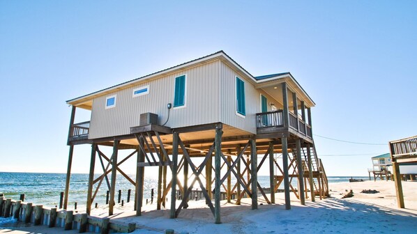 A wooden beach house with a balcony, built on stilts over the sand, near the ocean.