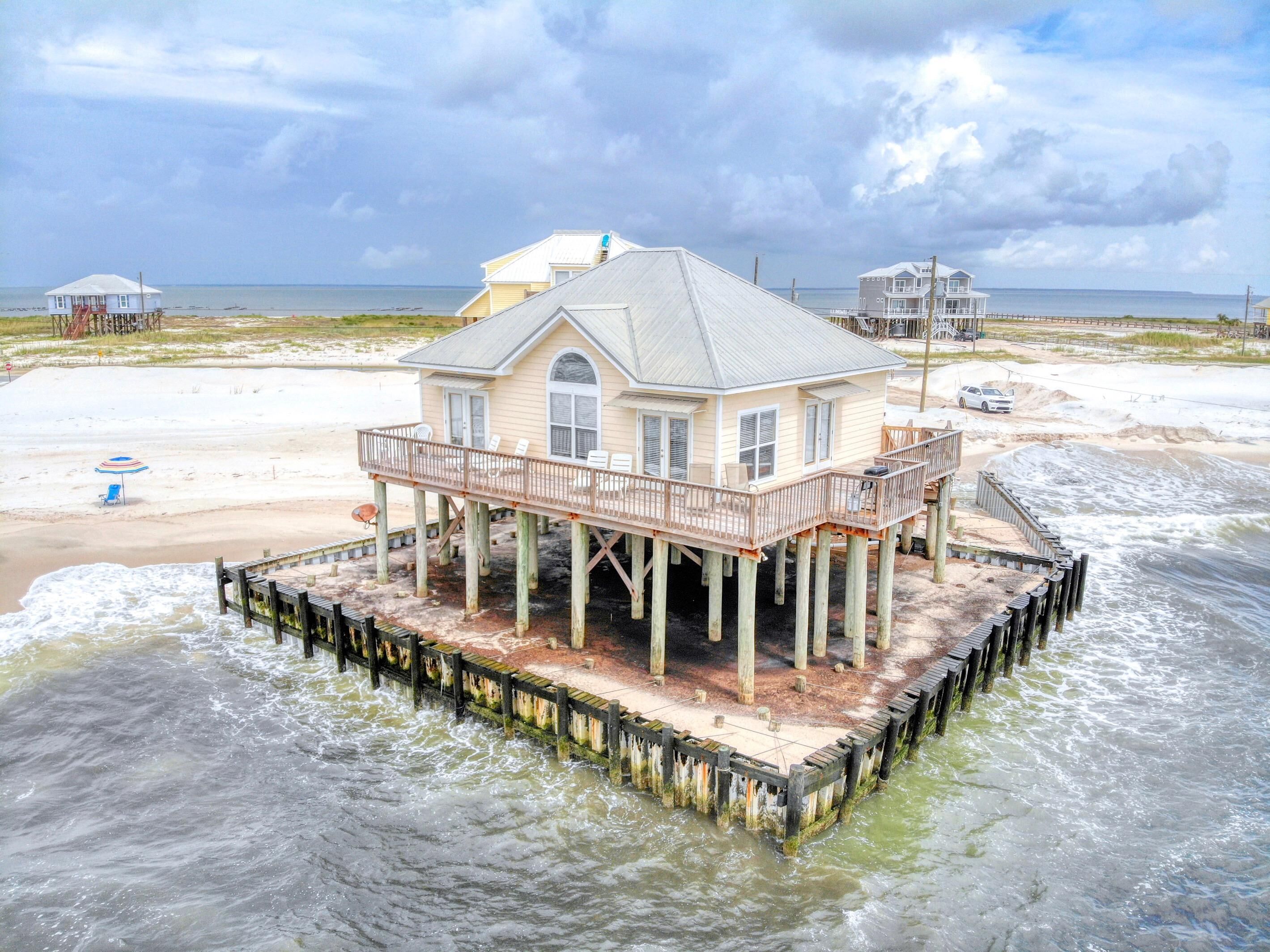Una casa su palafitte sopra l'acqua, con un pontile, una spiaggia sullo sfondo e un cielo nuvoloso.