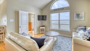 A spacious living room with a large window overlooking the beach, a flat-screen TV mounted on the wall, and a cozy seating area with a patterned rug. - Villa Delphinus - 103 Vargus C (Dauphin Island)
