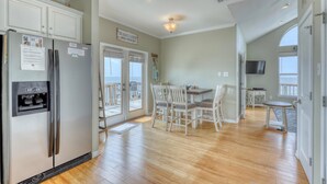 A spacious kitchen with a refrigerator, dining table, and chairs. There is a view of the ocean through the sliding glass doors. - Villa Delphinus - 103 Vargus C (Dauphin Island)