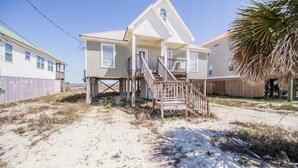 A beachfront house with a wooden staircase leading to a porch, a sandy beach in the foreground, and a clear blue sky. - Tropical Paradise - 2354 Islan (Dauphin Island)