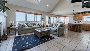 A spacious living room with a high ceiling, large windows, and a kitchen area with wooden cabinets and a bar stool. - Tropical Paradise - 2354 Islan (Dauphin Island)