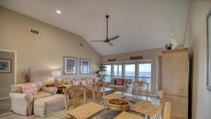 A spacious living room with a high ceiling, a ceiling fan, and a large window overlooking the ocean. There are two sofas, a coffee table, and a dining table with placemats. - Rainbows End - 100-b Shell Ct (Dauphin Island)