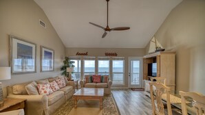 A spacious living room with a beige sofa, a wooden coffee table, and a dining area with wooden chairs. There is a ceiling fan, framed artwork on the walls, and a view of the beach through large windows. - Rainbows End - 100-b Shell Ct (Dauphin Island)