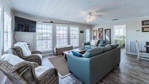 A spacious living room with a large sofa, two wicker chairs, a ceiling fan, and a television mounted on the wall. - Sea Spot - 2252 Mallard (Dauphin Island)