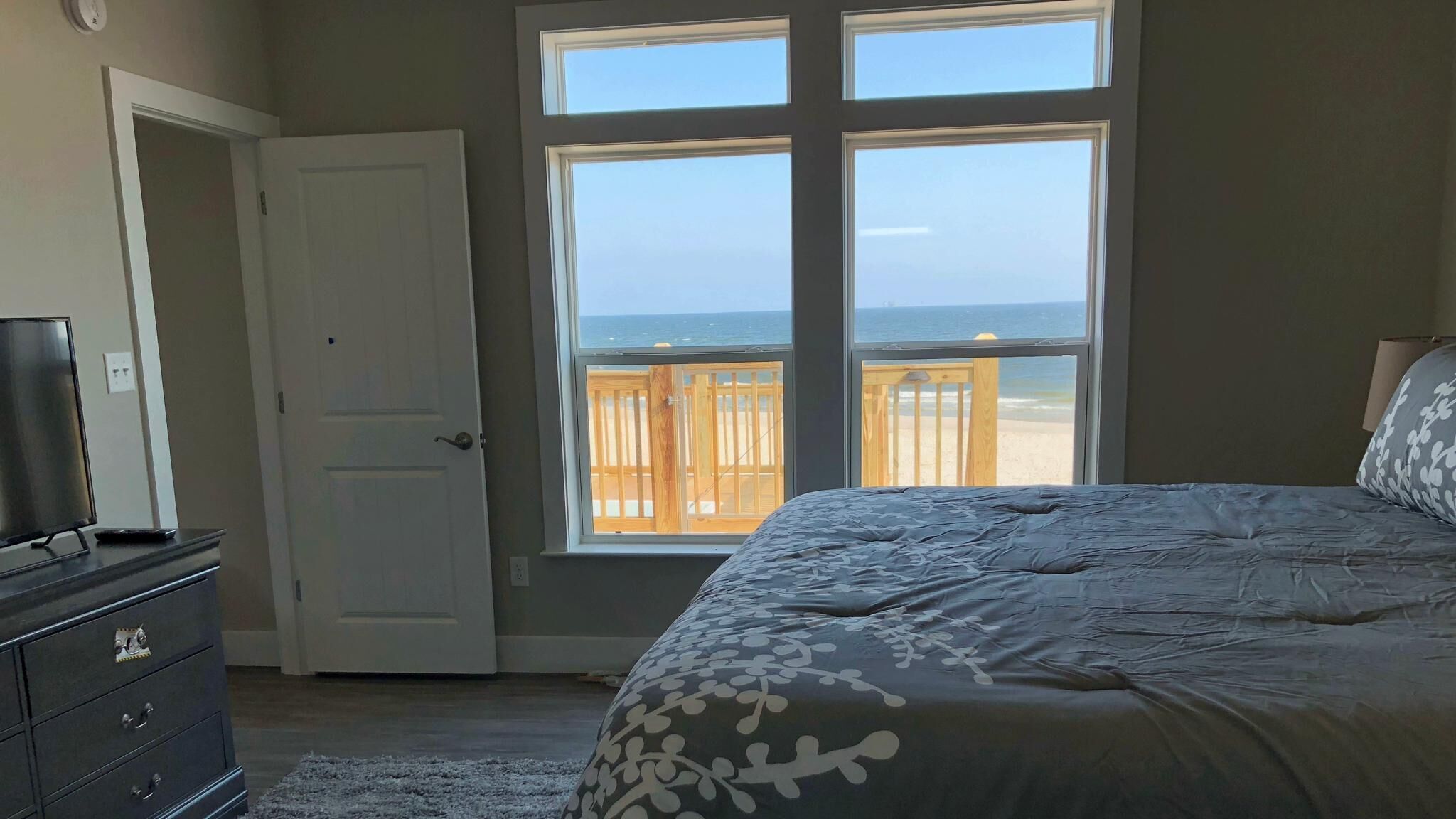 Un dormitorio con un ventanal grande con vista a la playa, una cama con colcha estampada, un aparador y un televisor sobre un mueble.