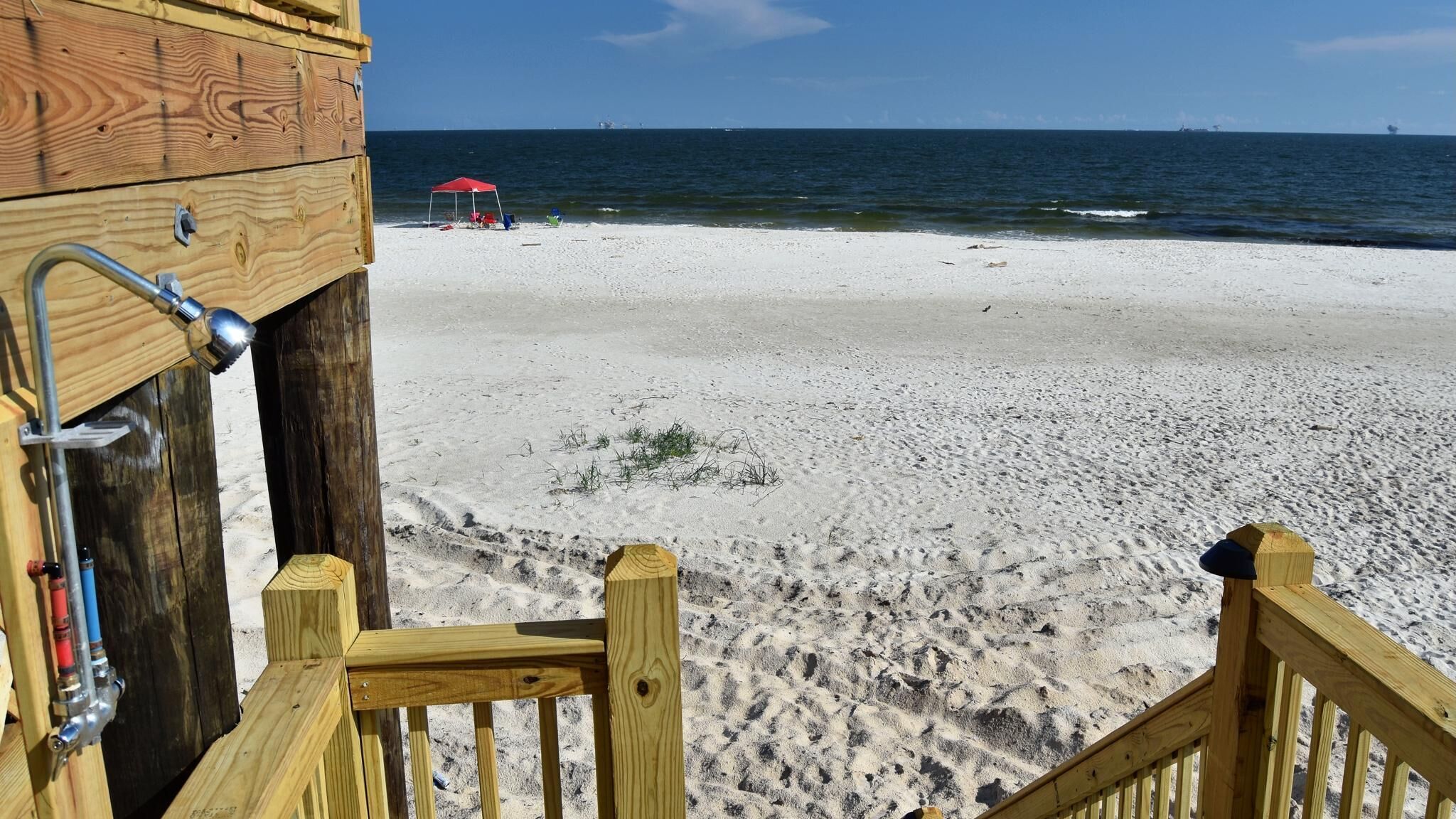 Une structure en bois installée sur la plage, avec une rambarde en métal, un parasol rouge et une plage de sable où l’on peut voir des empreintes.