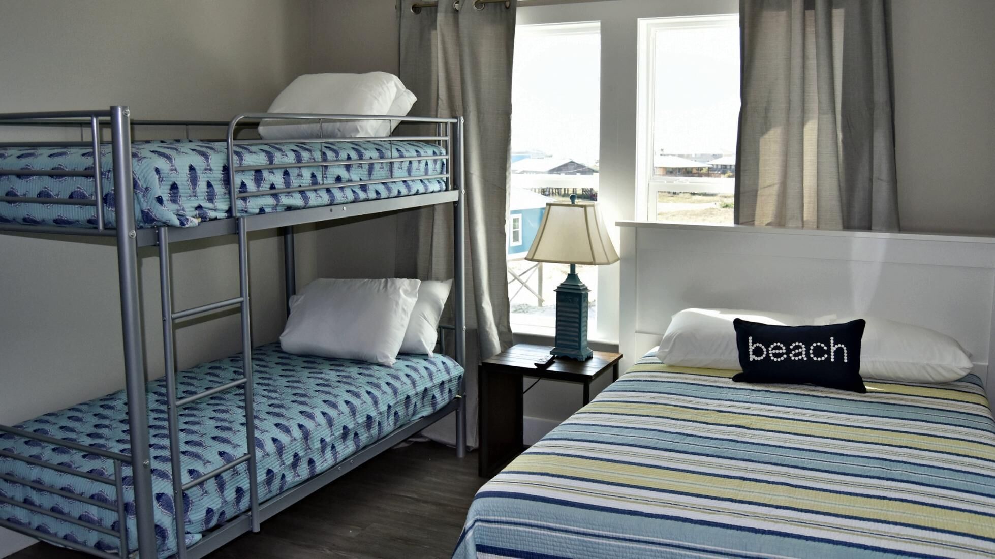 Una habitación con literas, una mesita de noche con lámpara, una cama con colcha a rayas y una almohada con la palabra 'beach'.